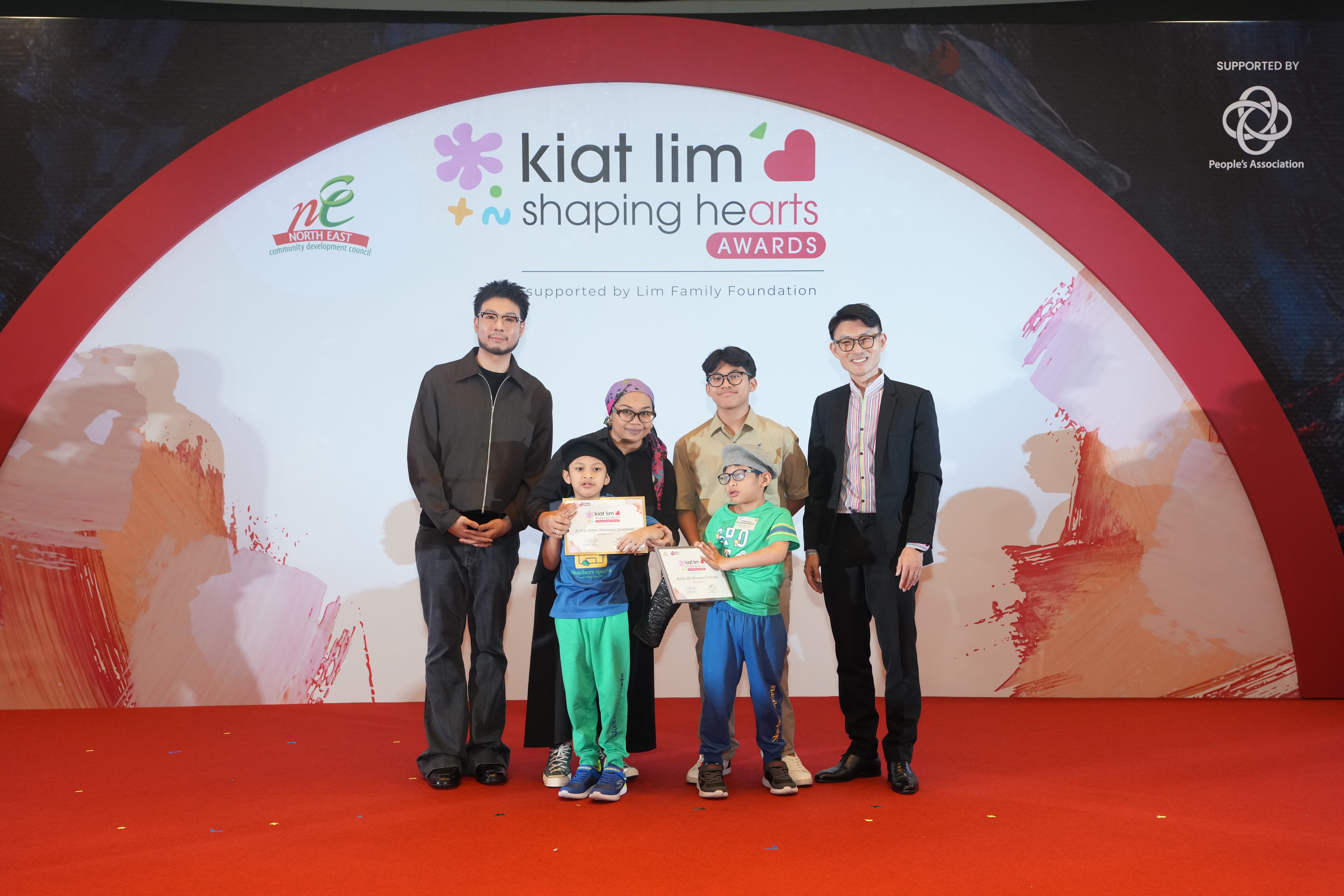 Award recipient standing on stage with the presenter, holding their certificate and posing for a group photo during the award ceremony, with a backdrop reading ‘Kiat Lim-Shaping Hearts Awards'.